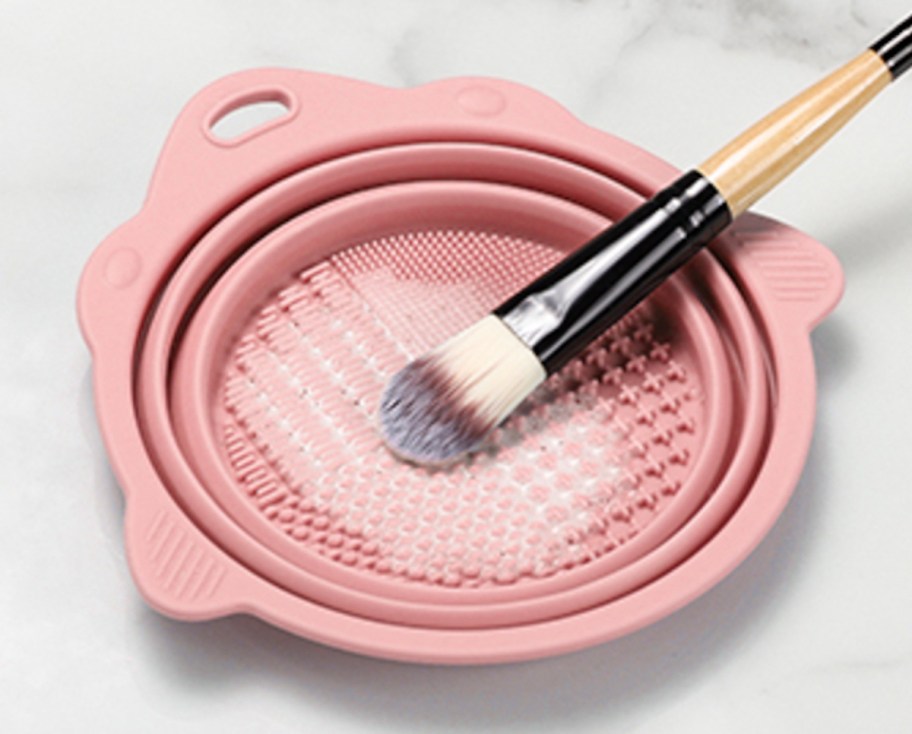 Makeup brush with soap in bottom of pink silicone cleaning bowl