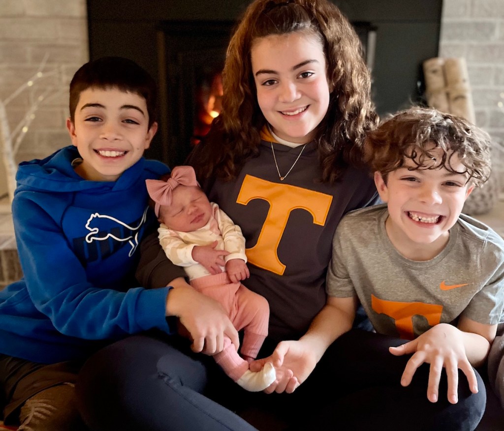 kids holding newborn baby by fireplace