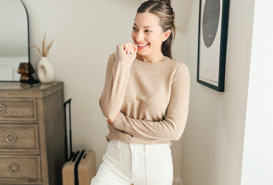 woman posing in beige sweater in bedroom with dresser and luggage