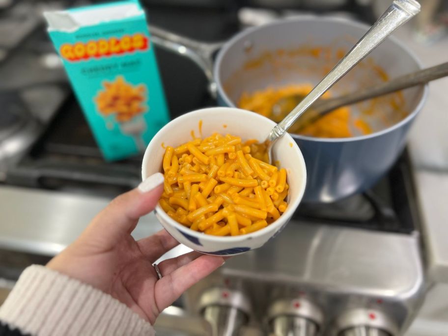 bowl of Goodles mac and cheese in person's hand with stove, pot and box in background