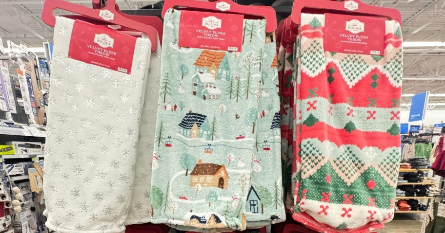 3 plush throw blankets on a store display, one white with snowflakes, one light blue with winter homes and trees, and one red, green, and white sweater print