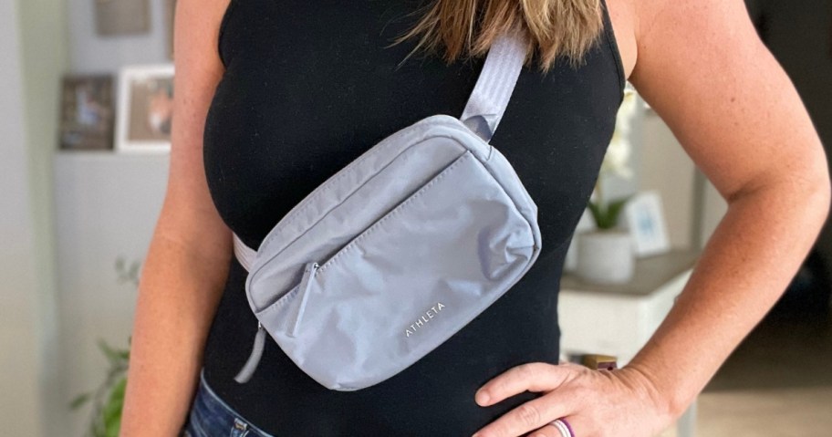 woman wearing a black sleeveless top, hand on her hip, carrying a grey belt bag as a crossbody bag