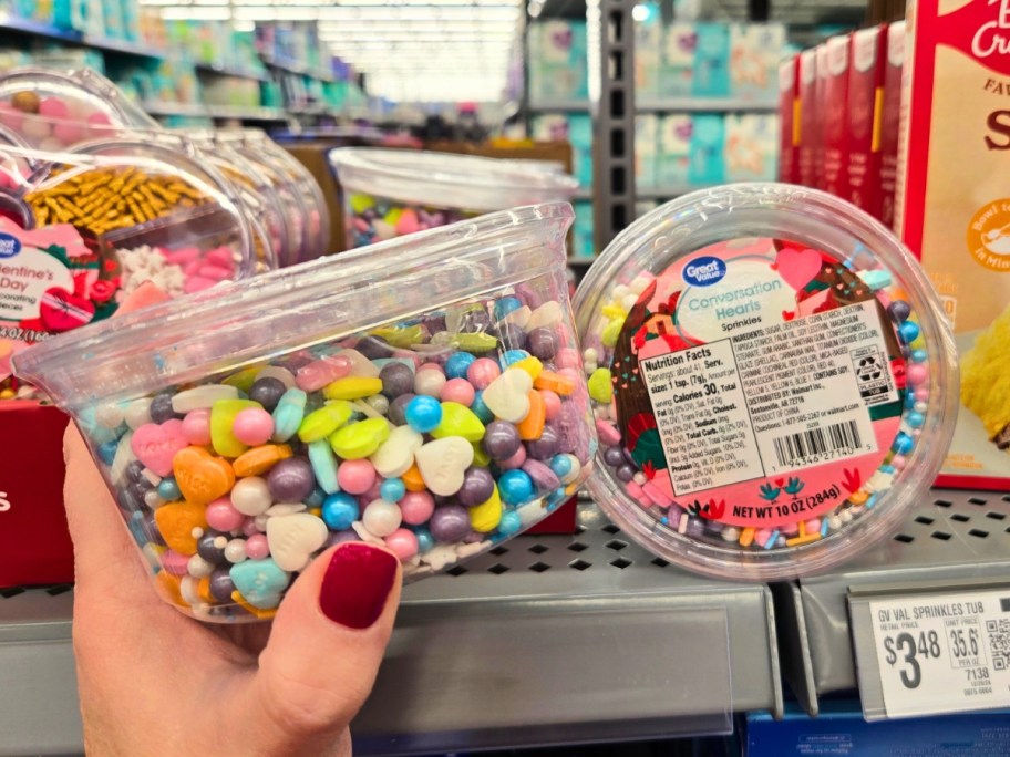 hand holding a clear plastic container with colorful Valentine's sprinkles in it, more containers on a store shelf behind it