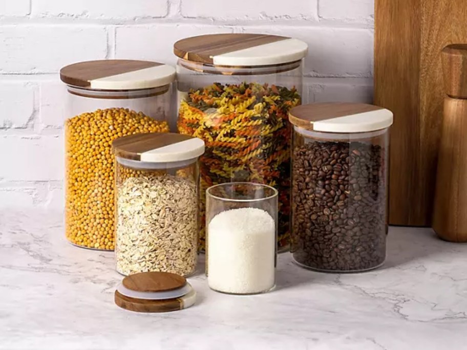 set of 5 glass canisters with acacia wood and marble tops filled with food on a kitchen counter