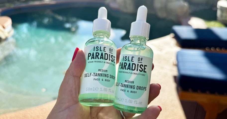 hand holding two green bottles of Isle of Paradise Self-Tanning Drops with pool in background