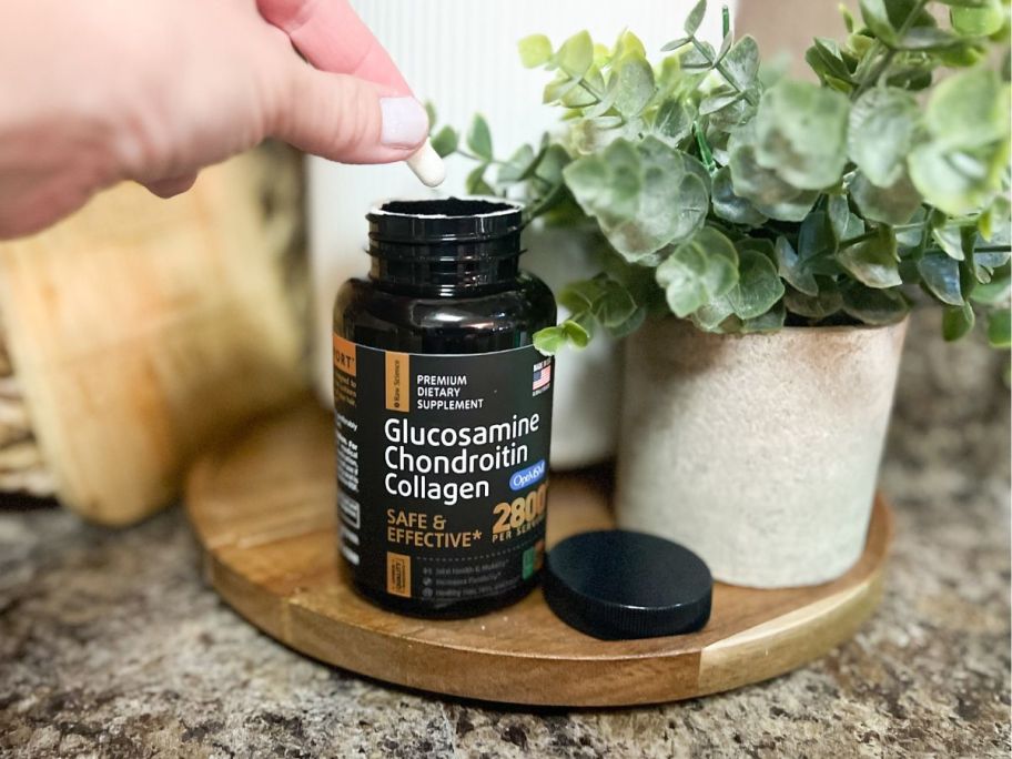 hand pulling a supplement out of a bottle of Raw Science supplements that's sitting on a tray beside a plant