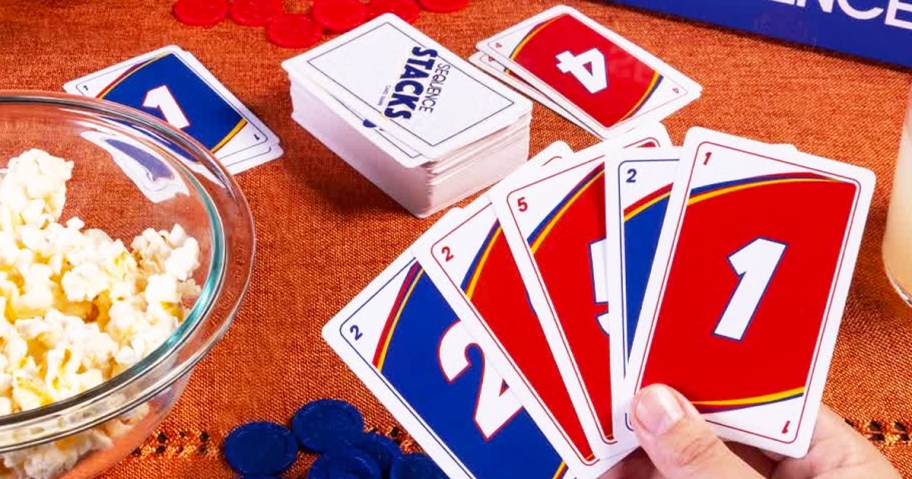 person holding sequence stacks card game cards with more cards and bowl of popcorn in background