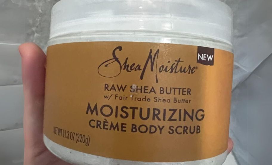 a womans hand holding a jar of SheaMoisture raw shea butter body scrub 