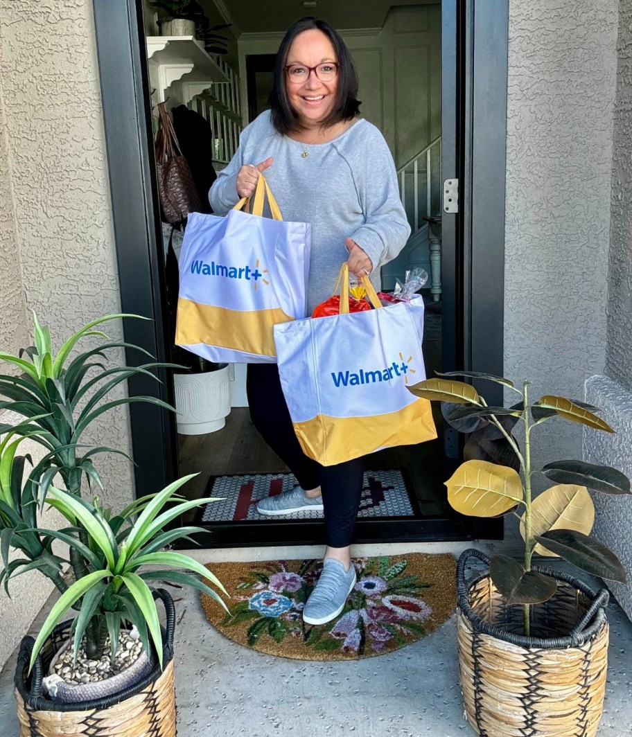 smiling woman standing on her doorstep with the door open, holding two walmart plus grocery delivery bags