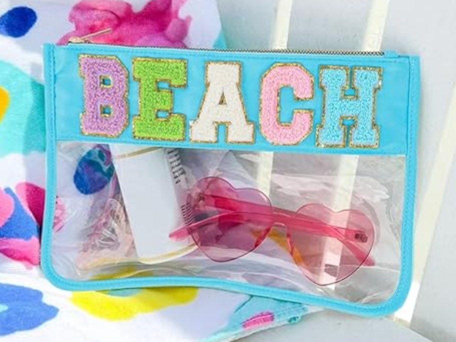 clear and blue zipper bag with fuzzy block letters that says BEACH filled with beach items on a chair with a beach towel