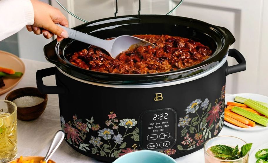 a slow cooker shown with chili cooking and a womans hand stirring with a spoon