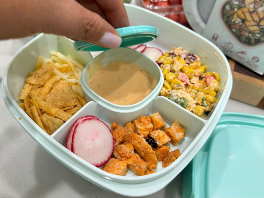 A Bentgo salad container with all of the onlinepartments filled with salad ingredients and a hand lifting the lid off the dressing onlinepartment in the middle