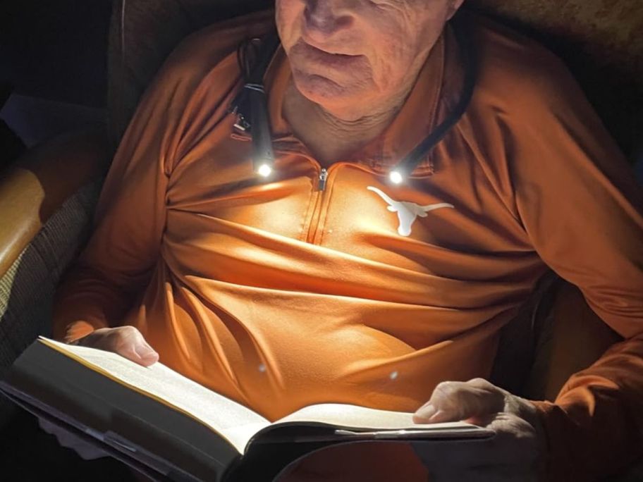 man wearing an LED neck reading light reading a book sitting in a chair in a dark room