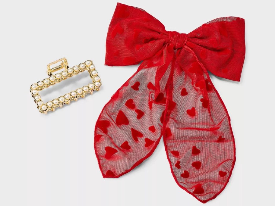 a gold hair claw clip with pearls next to a red hairbow with red hearts on it