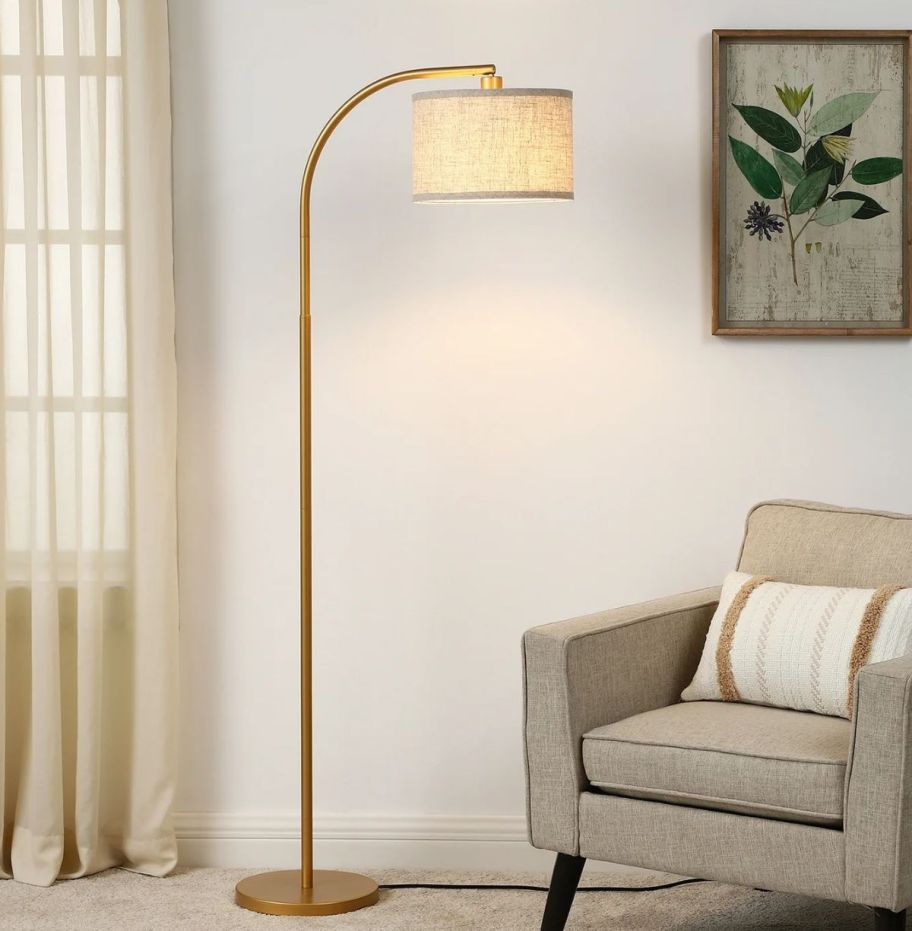 gold floor lamp with curved arm pictured next to a chair adjacent to a window