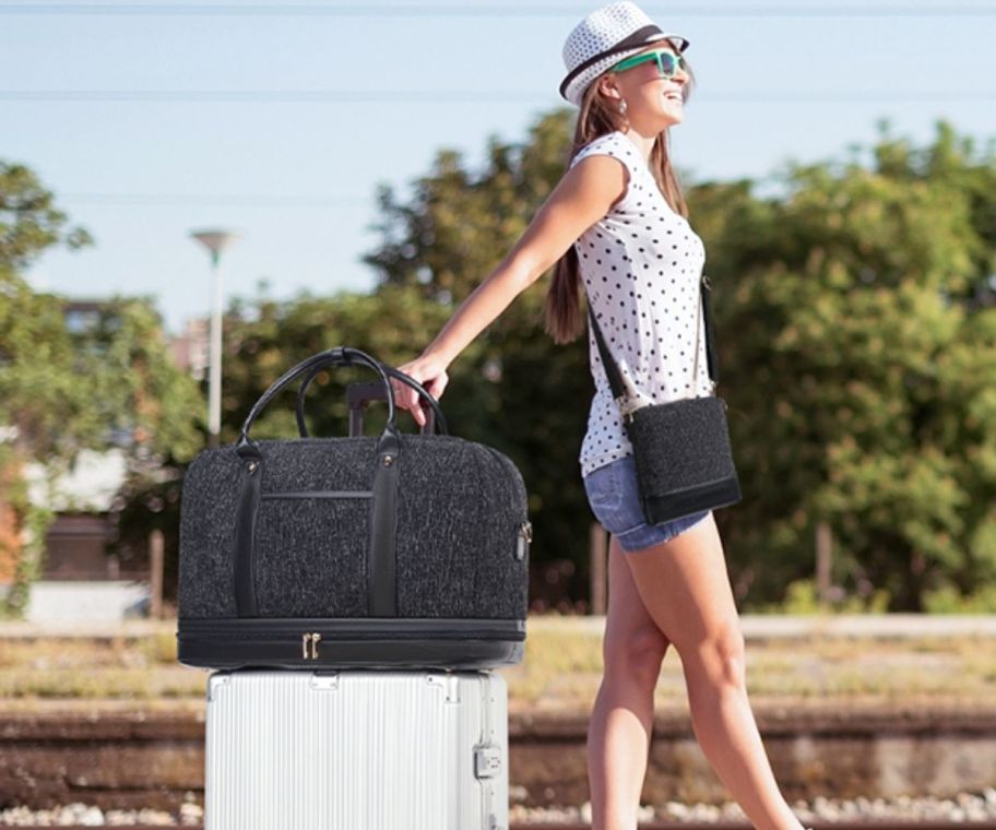 a woman with a travel duffle on top of a rolling suitcase, standing buy a train track