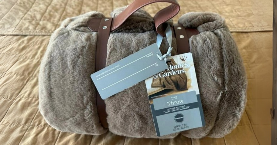 a greyish taupe color faux fur blanket with a faux leather double strap around it and tags that say "Better Homes and Gardens"