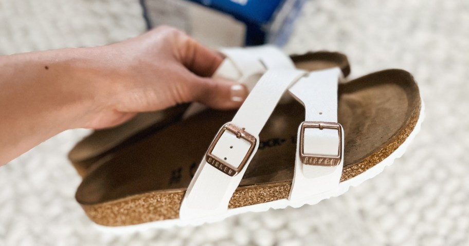 hand holding a pair of white and brown Birkenstock slide sandals, shoe box on the floor in the background