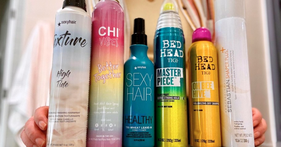 various hairspray cans from Chi, Bed Head, Sexy Hair, and Sebastian on a bathroom counter, person's hands on each side