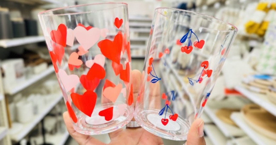 a hand holding up 2 clear drinking glasses, one has red and pink hearts and one has red and blue cherry hearts