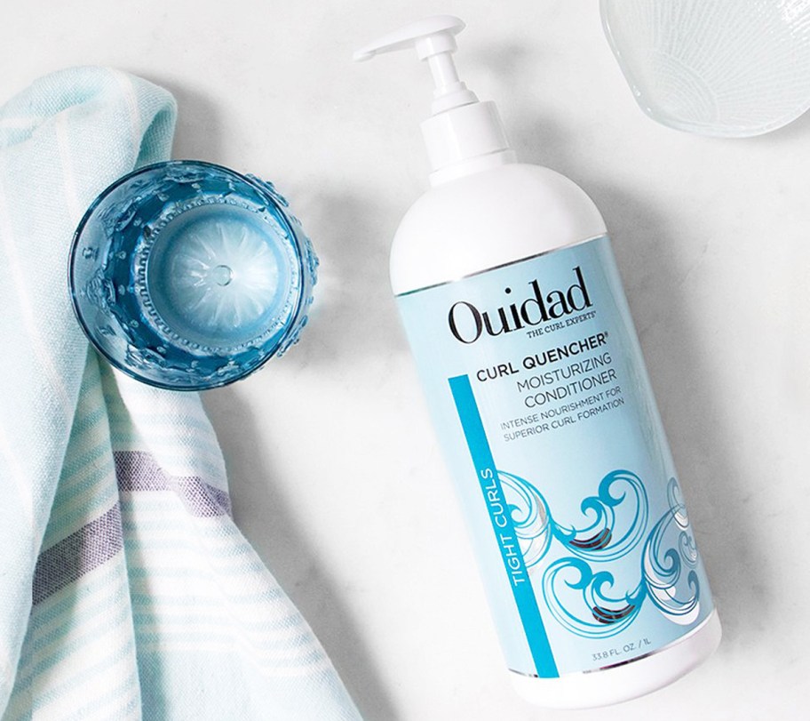 large blue and white bottle of Ouidad Curl Quencher Moisturizing Conditioner near glass of water and towel