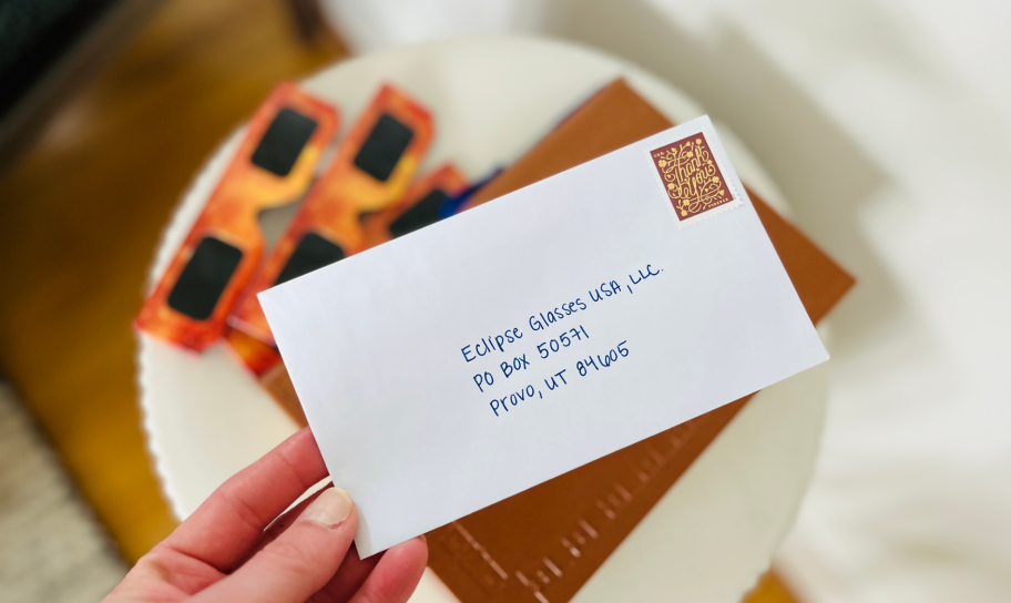 hand holding envelope with address and stamp on it with eclipse glasses in background on a table