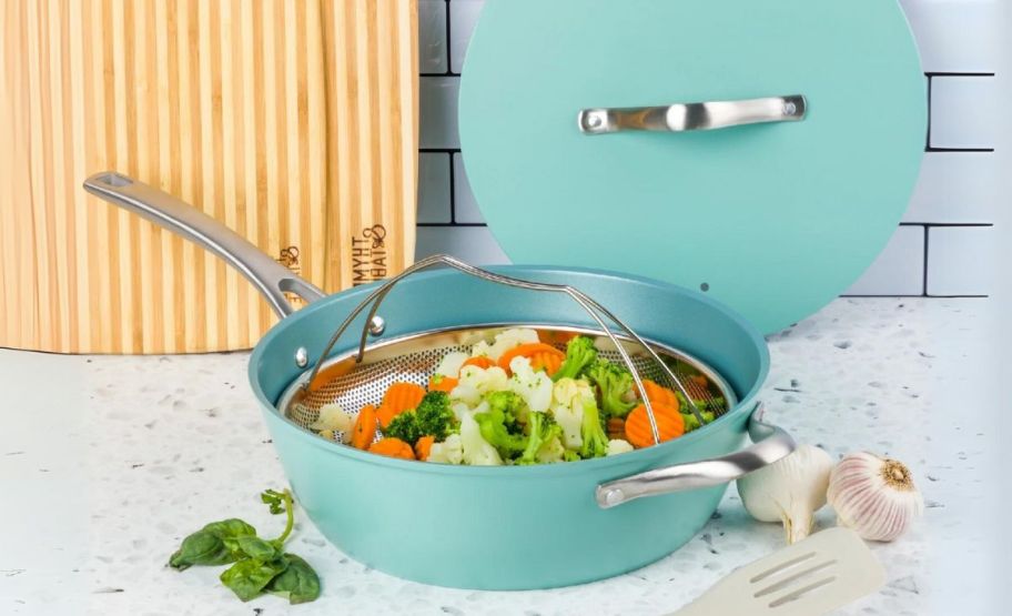 a turquoise blue non-stick chefs pan, lid and steamer basket shown with steamed vegetables and sitting on a kitchen counter