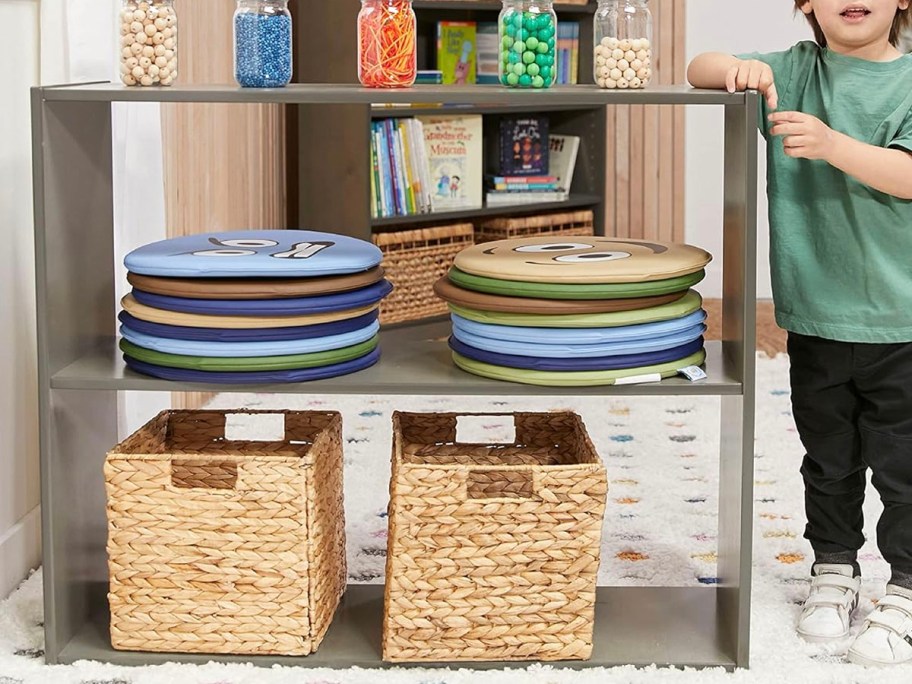 boy standing next to brown 2 shelf cabinet with toys and baskets on it