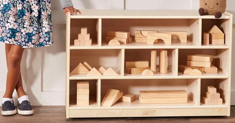 child stanidng next to wood cabinet full of wooden building toys