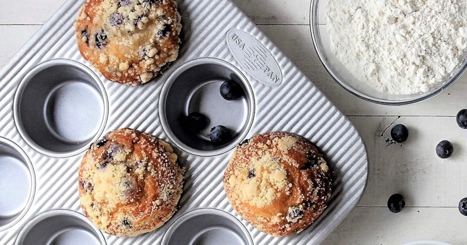 blueberry muffins and blueberries in muffin pan next to a bowl of flour