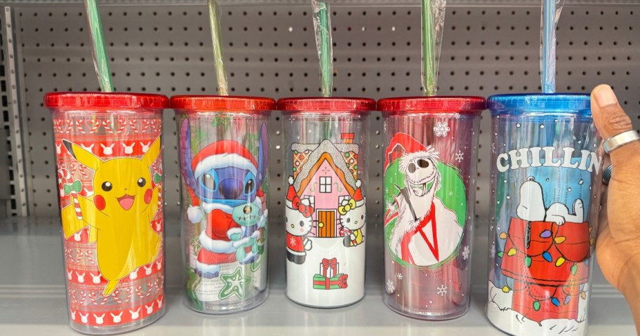 plastic tumblers with straws and lids that have characters in Christmas styles on them - Pikachu, Stitch, Jack Skellington, Hello Kitty and Snoopy - on a store shelf, hand reaching for the Snoopy one