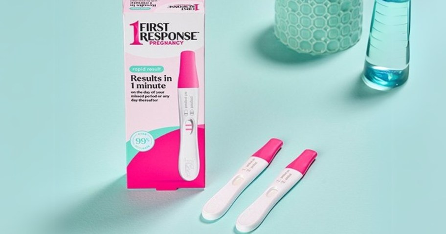 a pink and white First Response Pregnancy test box with 2 pink and white tests laying beside it on a green background