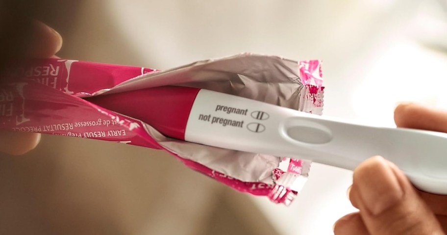 hand holding a pink and white pregnancy test, pulling it out of the wrapper it onlinees in