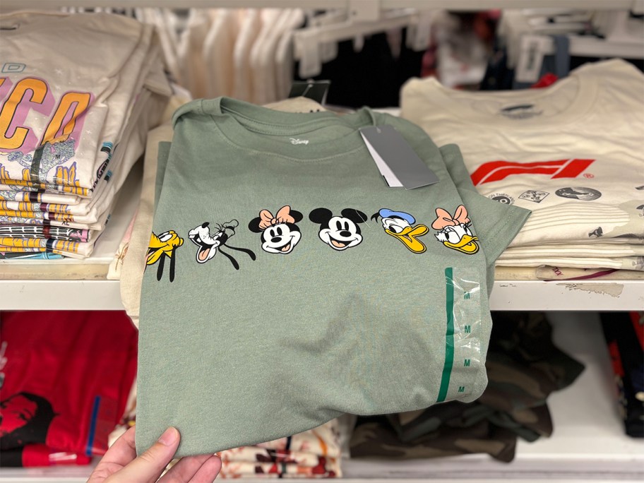 Green Disney Mickey and Friends graphic T-shirt at Target featuring Pluto, Goofy, Minnie, Mickey, Donald, and Daisy, displayed on a store shelf.