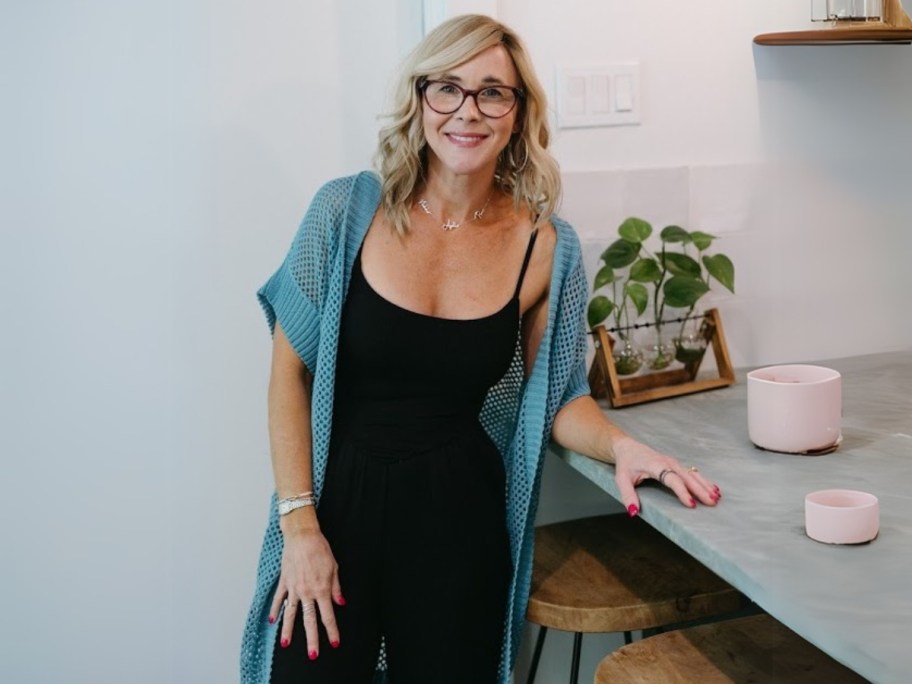 Collin wearing a blue crochet duster with a black bodysuit and pants standing next to a kitchen counter