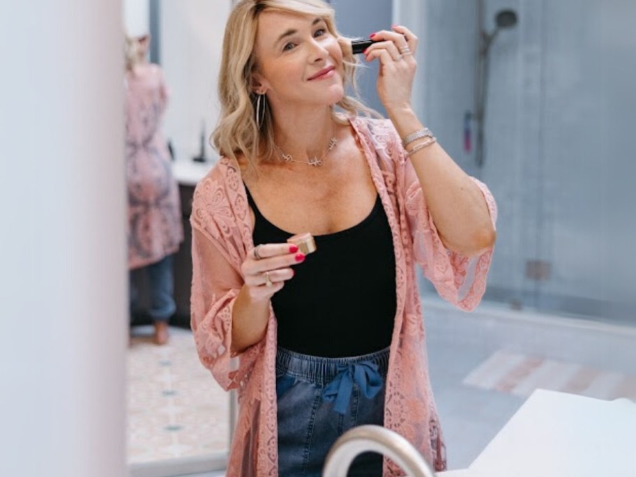 Collin putting on makeup in a mirror wearing a pink lace crochet duster, black body suit and jeans
