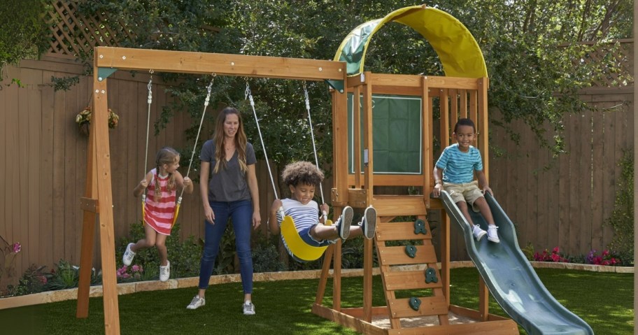 mom and kids playing on a wooden swingset with swings, slide, climbing wall and cover over the slide top