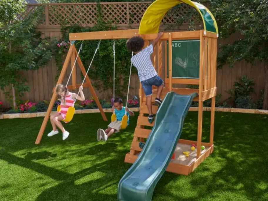  kids playing on a wooden swingset with swings, slide, climbing wall and cover over the slide top