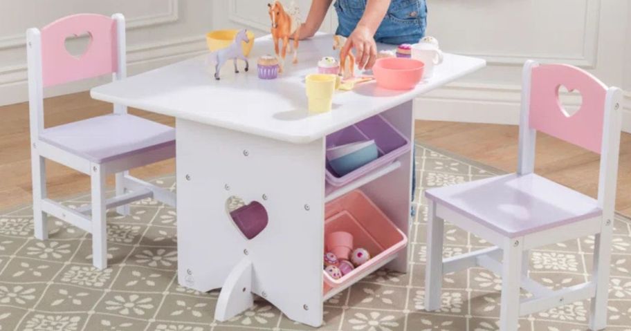 A kid's white and pink table and chair set with toys on top and a little girl playing with them