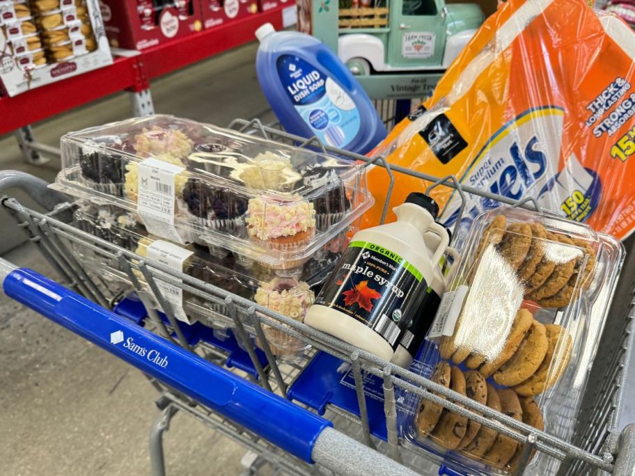 A sam's Club shopping cart filled with baked goods, maple syrup, paper towels and dish soap