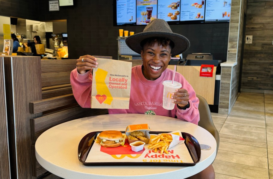 smiling woman holding a mcdonalds bag and drink with food on a tray in front of her