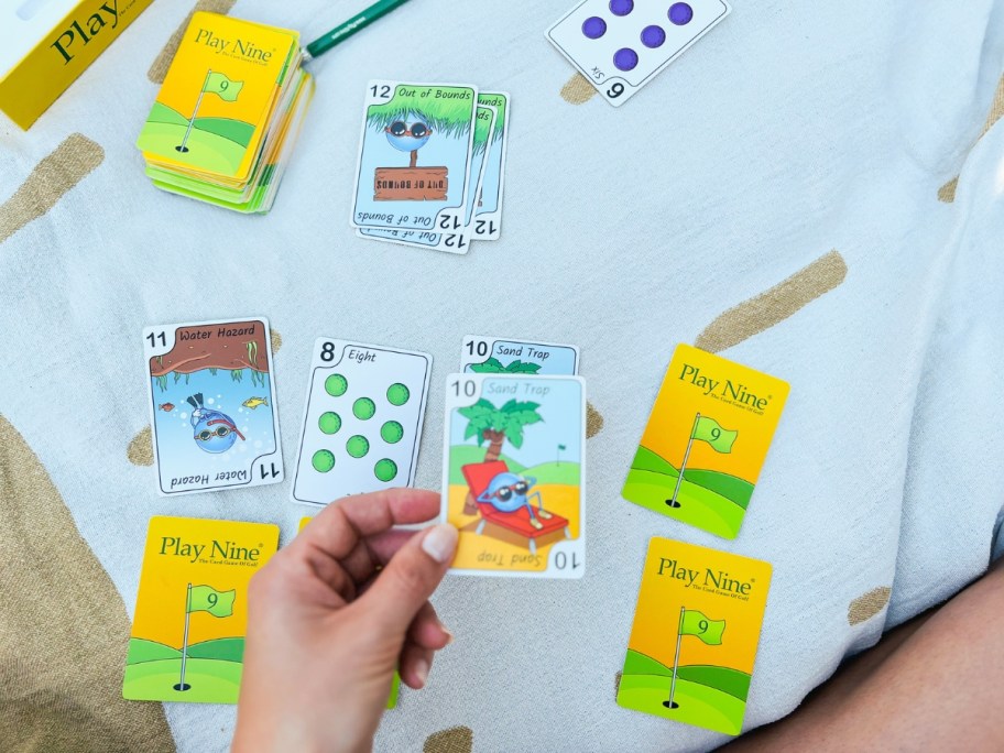 hand holding a card from a Play Nine Card game with more cards, score pad and pencil on a blanket outdoors