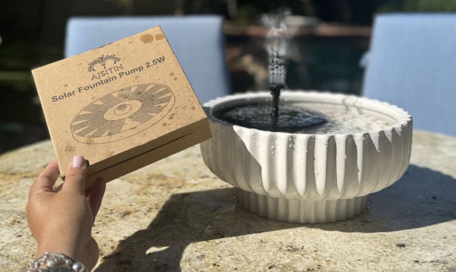 a solar tabletop fountain in a planter next to a womans hand holding the product packaging