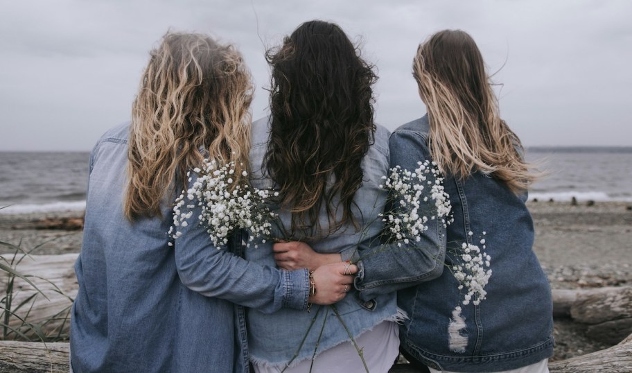 three women wearing jean jackets holding babies breath looking out at ocean