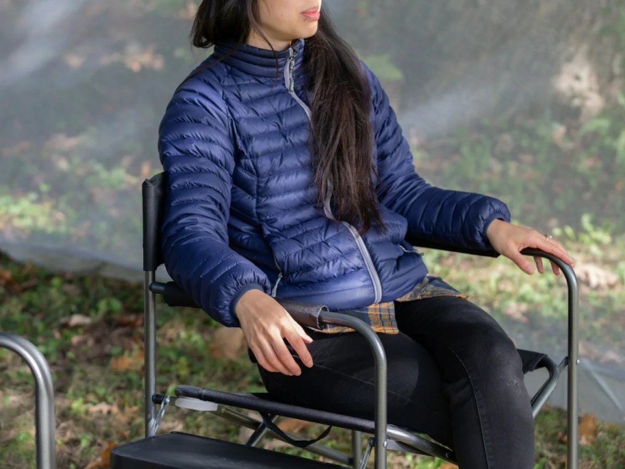 woman sitting in a black directors style camping chair with a side table outside by a tent