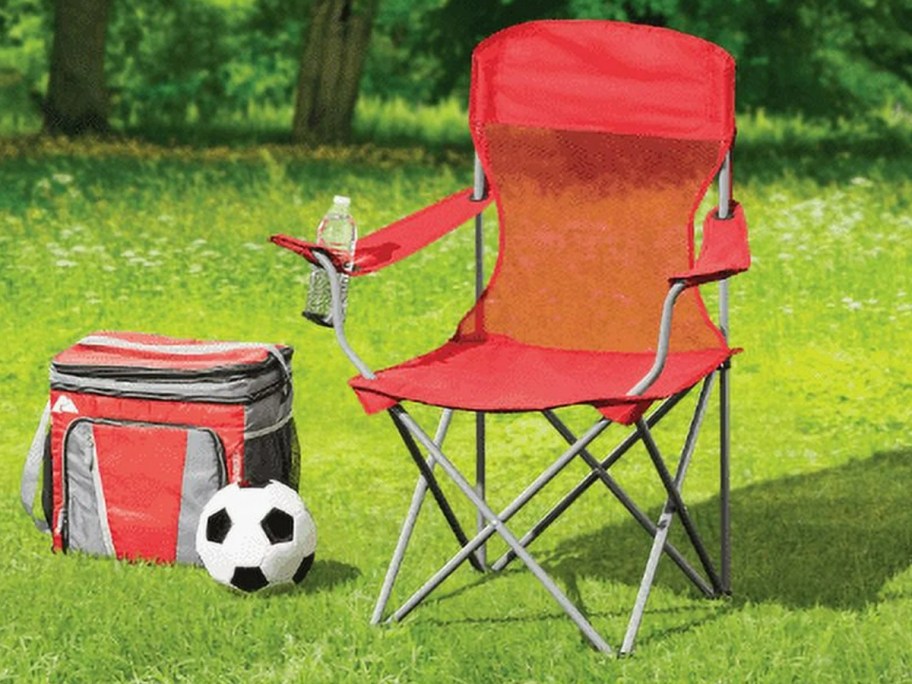 red mesh camping chair sitting outside on the grass, red cooler and soccer ball beside it