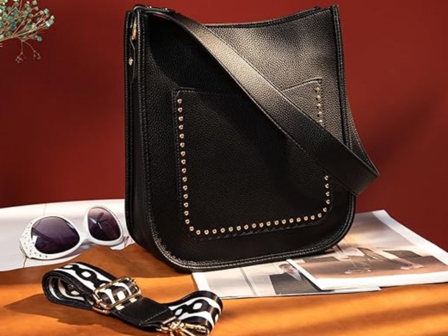 a black shoulder/crossbody pack with a front pocket with silver stud detail, extra guitar style pattern strap to the side - sitting on on a table with sunglasses, magazines, vase 