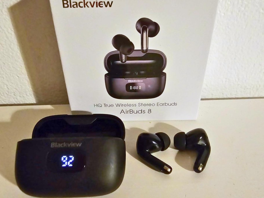 a pair of black wireless earbuds next to the charging case, a white box with the packaging they onlinee in behind them