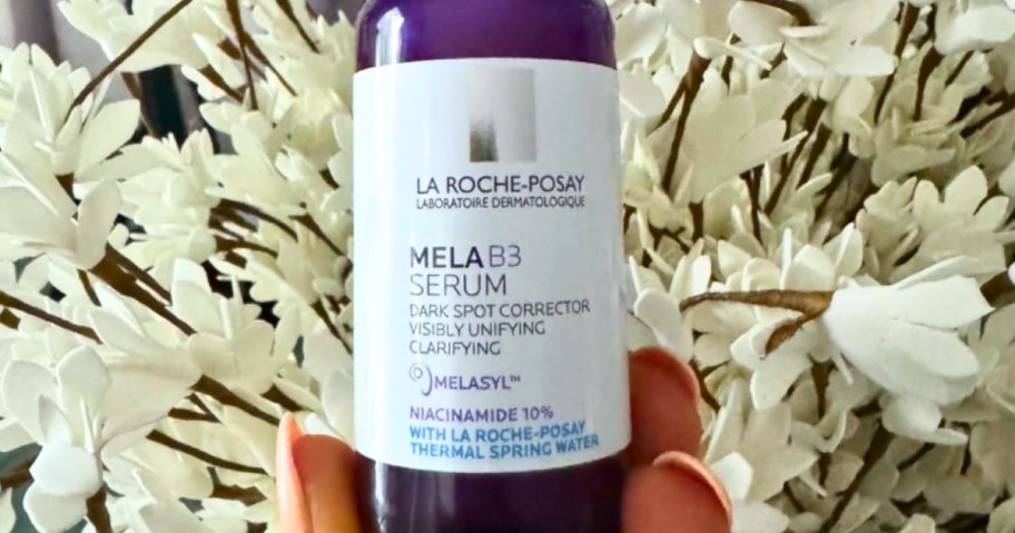 hand holding a small purple bottle with a white label of LaRoche Posay Mela B3 Dark Spot Serum, white flowers in the background