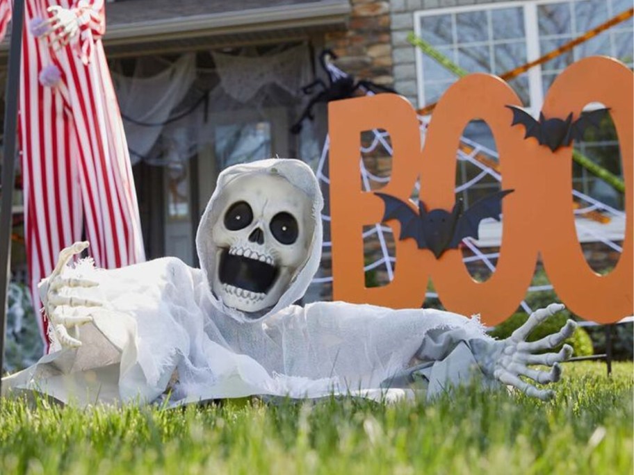 a yard with Halloween decor including a skeleton head, shoulders and arms onlineing out of the ground and a large orange sign that says "BOO" with black bats on it